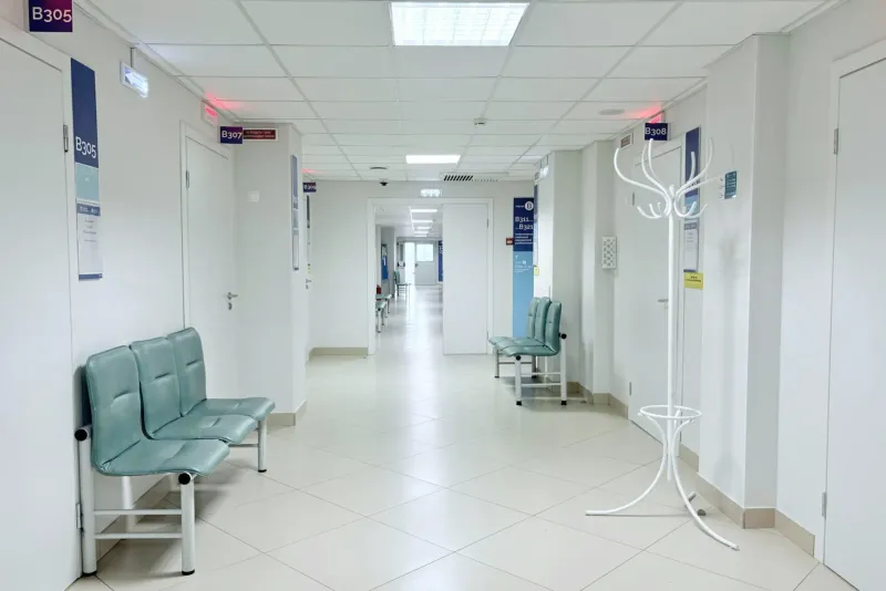 A bright hospital corridor with numbered doors and waiting chairs